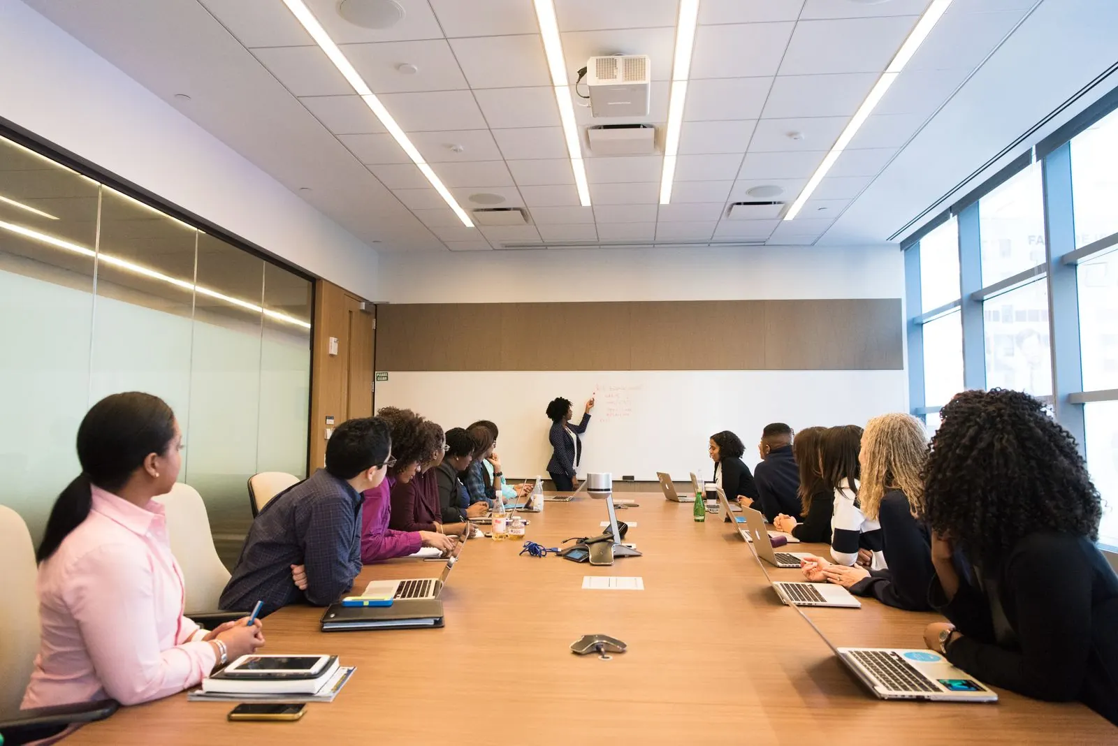 A facilitator at the front of a bright meeting room, with a team seated along a long table for a working session.